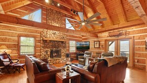 Ein Blockhaus-Innenraum mit Kamin aus Stein, Ledersofas, einem Fernseher und einem Deckenventilator.