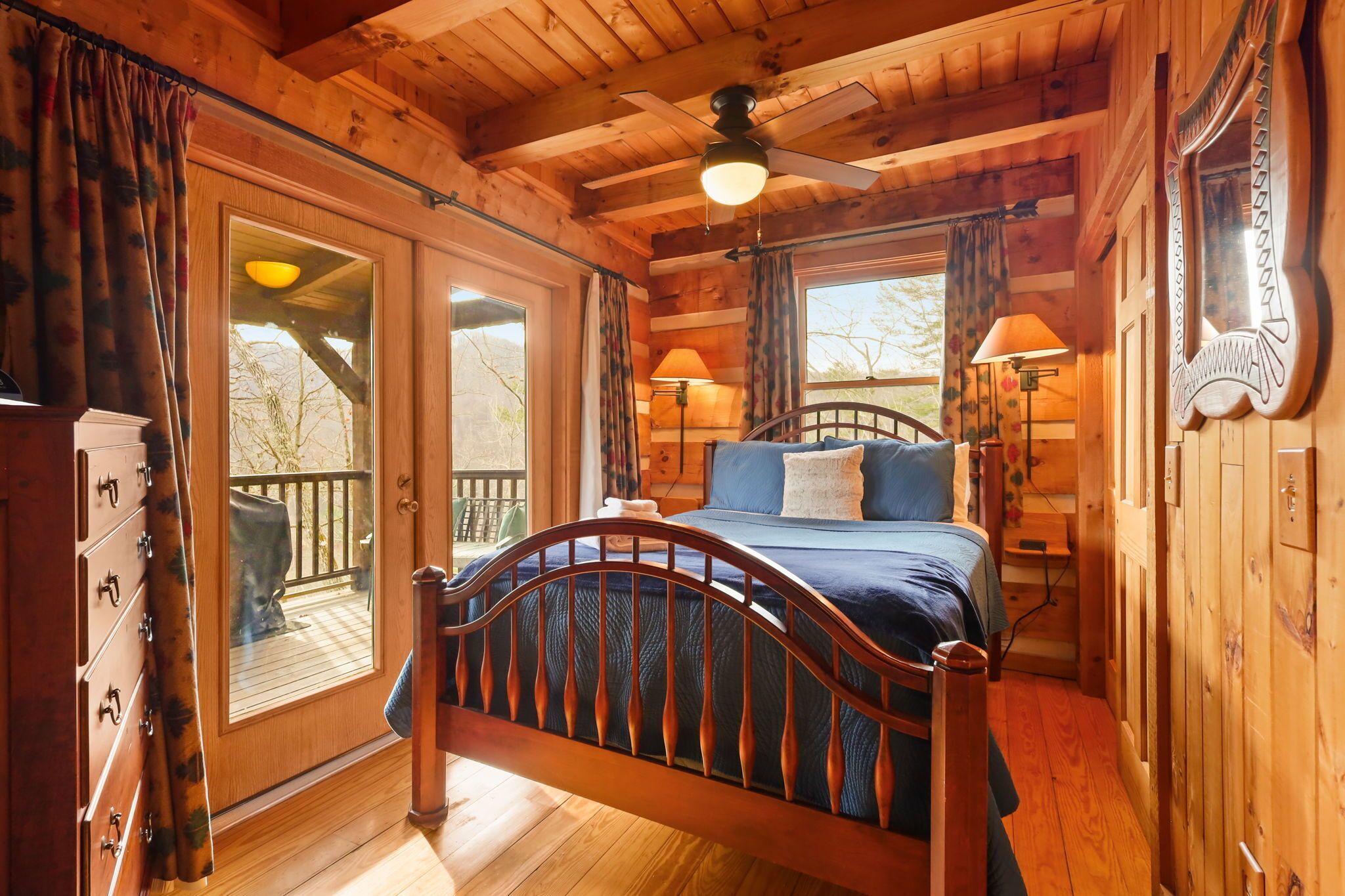 A wooden cabin bedroom with a bed, a ceiling fan, a window with curtains, and a view of the outside.