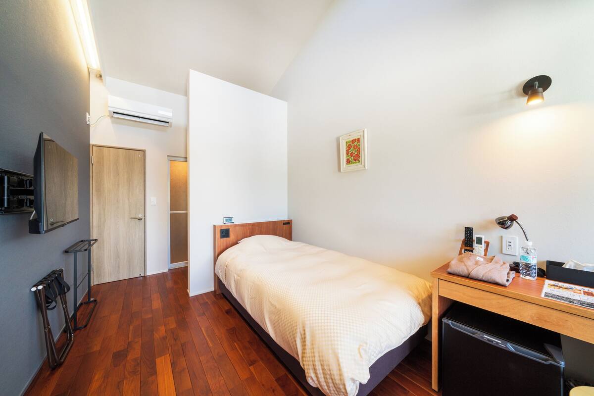 Un dormitorio pequeño y ordenado con suelo de madera, cama con colcha blanca, escritorio con lámpara y un refrigerador pequeño.
