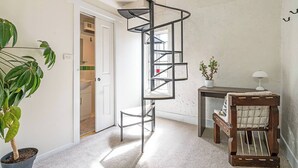 Una habitación pequeña con una escalera de caracol, una planta en maceta, una mesita con una lámpara y una silla de madera.
