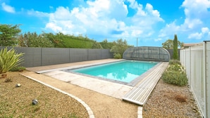 Una piscina rectangular con un deck alrededor, una zona cubierta y un jardín cercado.