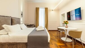 Ein Hotelzimmer mit Bett, Nachttisch, einem kleinen runden Tisch mit Stühlen, einem Fernseher an der Wand und einem Fenster mit Vorhängen.