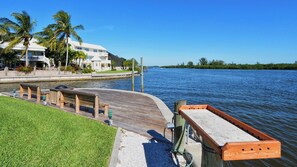 A waterfront property with a wooden dock, a grassy area, and a large house with a balcony overlooking the water.