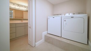 A laundry room with two white front-loading washers and dryers, a beige tiled floor, and a partially open door leading to a kitchen area with a countertop and cabinets. - Heated Pool, FREE WIFI Luxury Bayside Condo Across from Siesta Key Beach | Beautiful 3BR/2BA Gulf & Bay Club Bayside Condo (Sarasota)