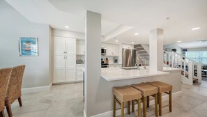 A modern kitchen with white cabinetry, a central island, and a dining area with wicker stools. - Heated Pool, FREE WIFI Luxury Bayside Condo Across from Siesta Key Beach |  Beautiful 3BR/2BA Gulf & Bay Club Bayside Condo (Sarasota)