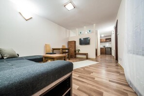 A modern, well-lit room with a sofa, a wooden dining table, a TV mounted on the wall, and a kitchen area in the background.