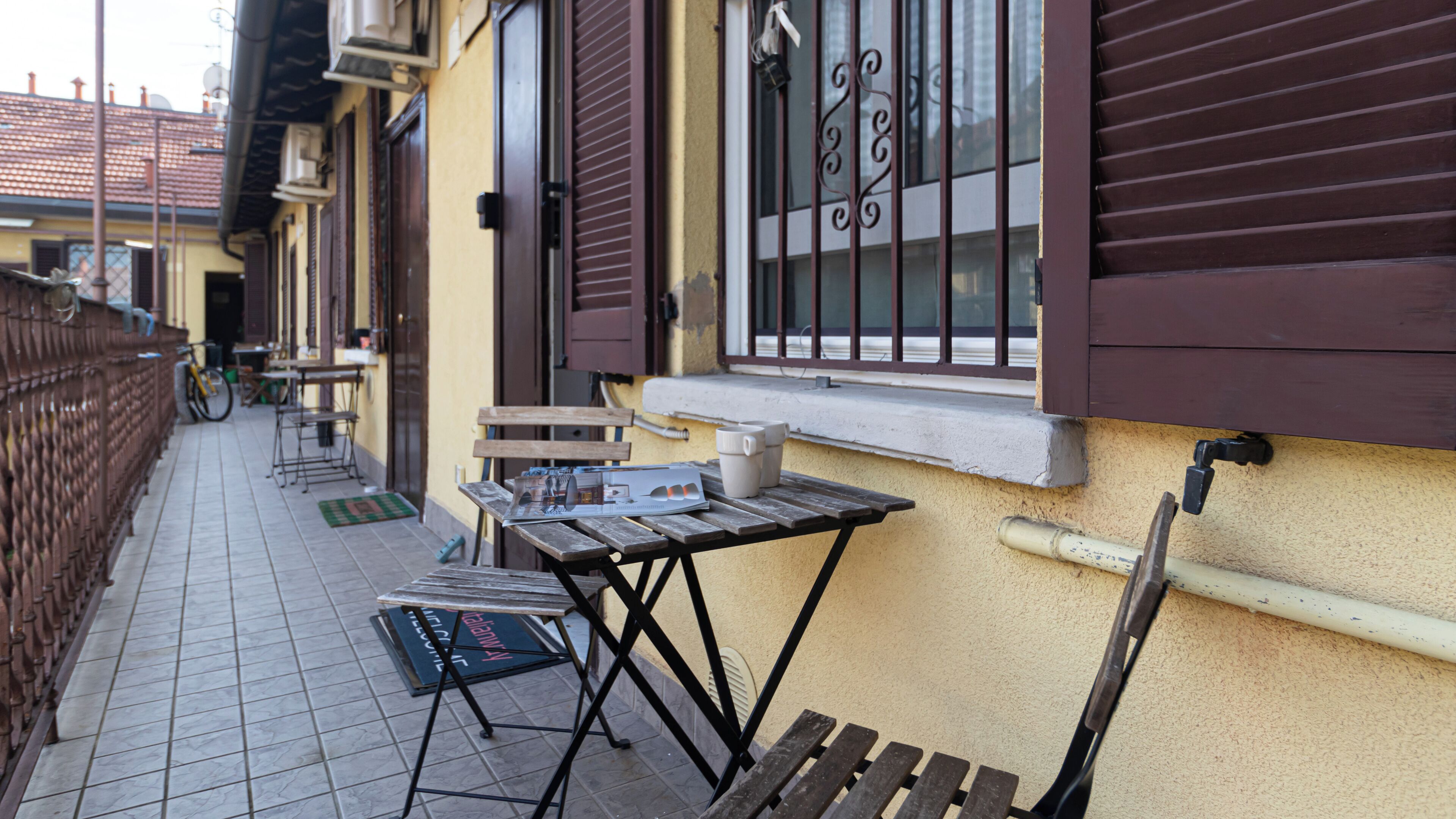 A narrow outdoor corridor with a wooden bench, a table with a newspaper, and a bicycle.
