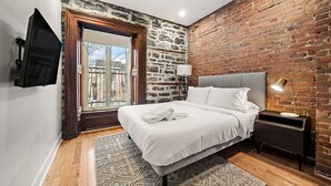 Ein modernes Schlafzimmer mit Backsteinwand, großem Fenster, Bett mit weißer Bettwäsche, Nachttisch und einem an der Wand befestigten Fernseher.
