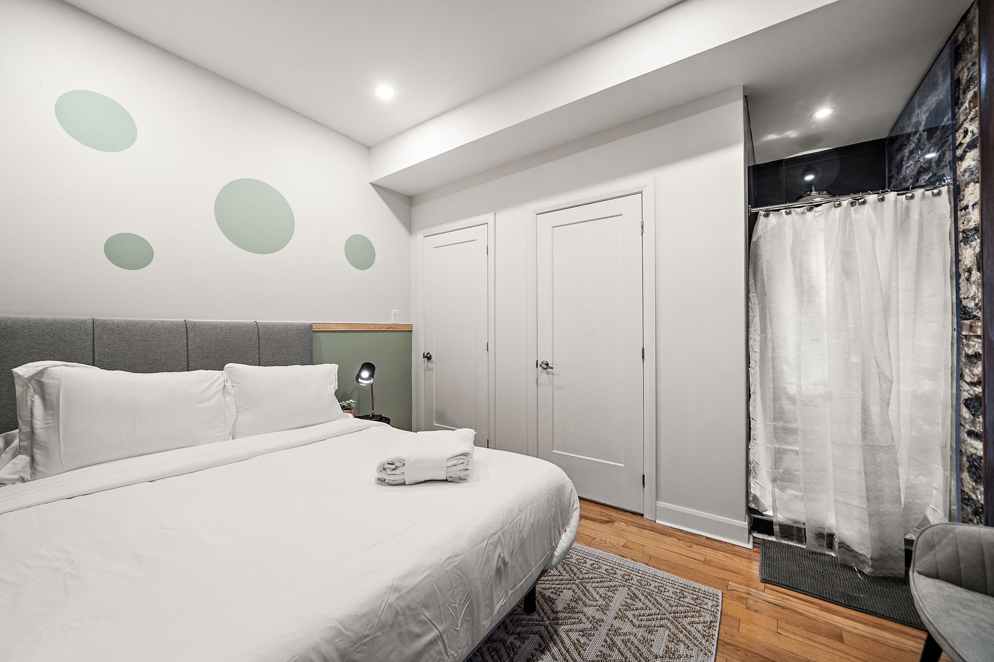 Un dormitorio moderno con una cama grande, un cabecero gris, una mesita de noche con una lámpara y una zona de ducha con cortina.