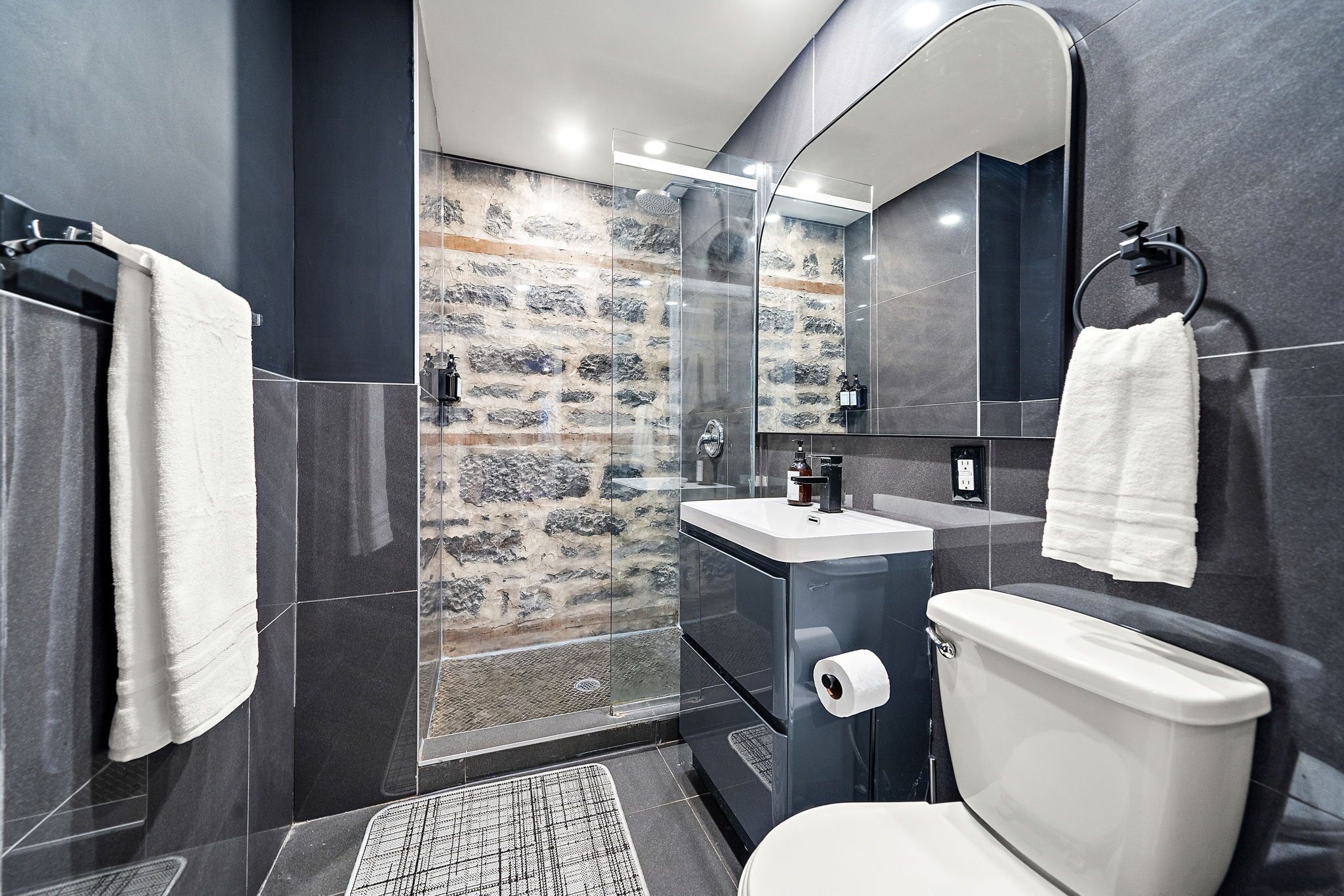 A modern bathroom with a stone wall, a glass shower enclosure, a white toilet, and a sink with a mirror above it.
