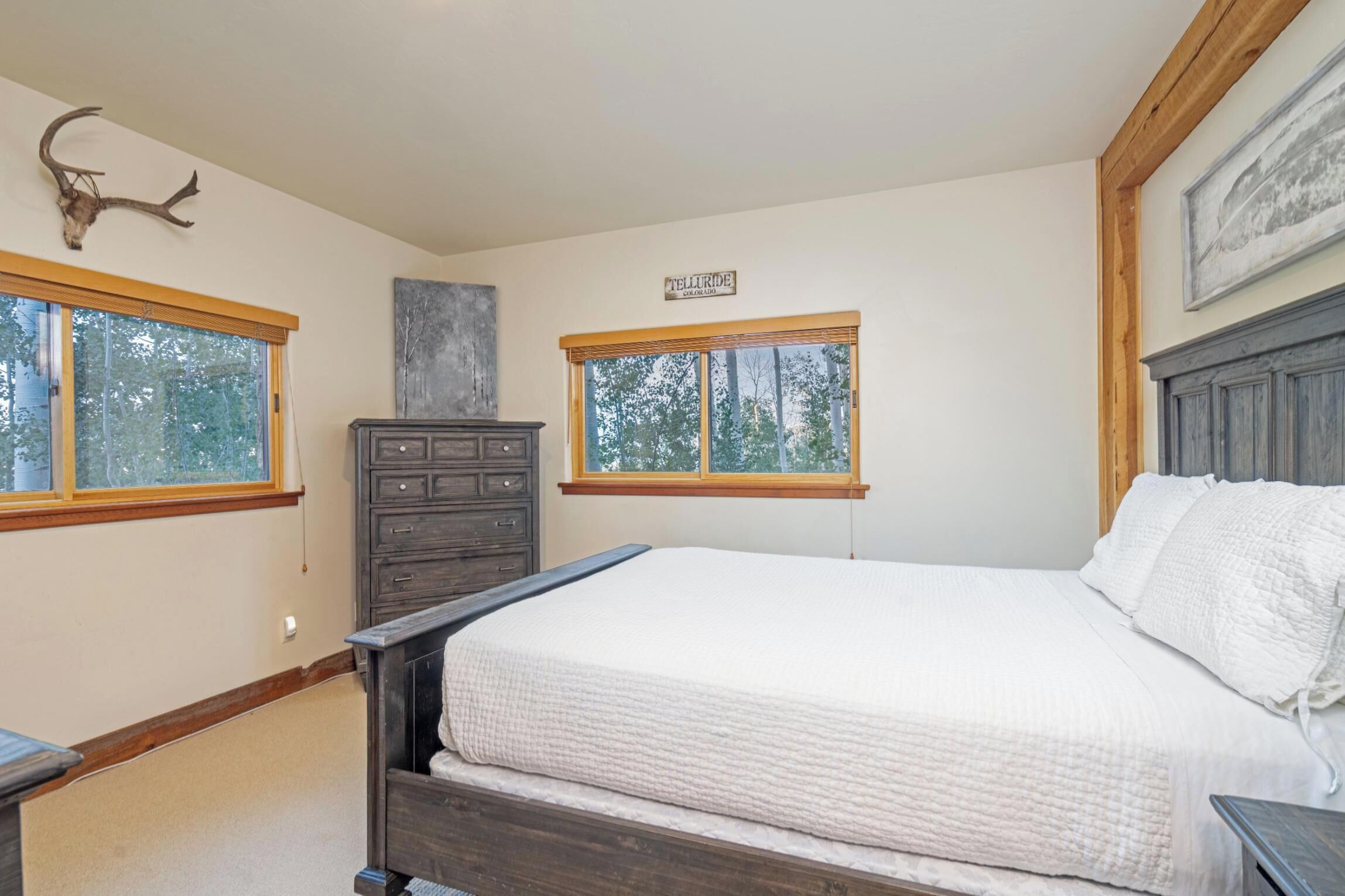 A bedroom with a large bed, two windows, a dresser, and a wall-mounted deer skull.