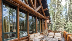 A log cabin with a deck featuring a sofa and tables, large windows, and a view of tall trees.