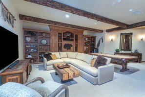 A living room with a sectional sofa, a pool table, wooden shelves, and a television.