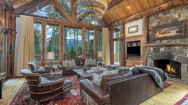 A rustic interior with a stone fireplace, leather sofas, and a large window overlooking a forest.