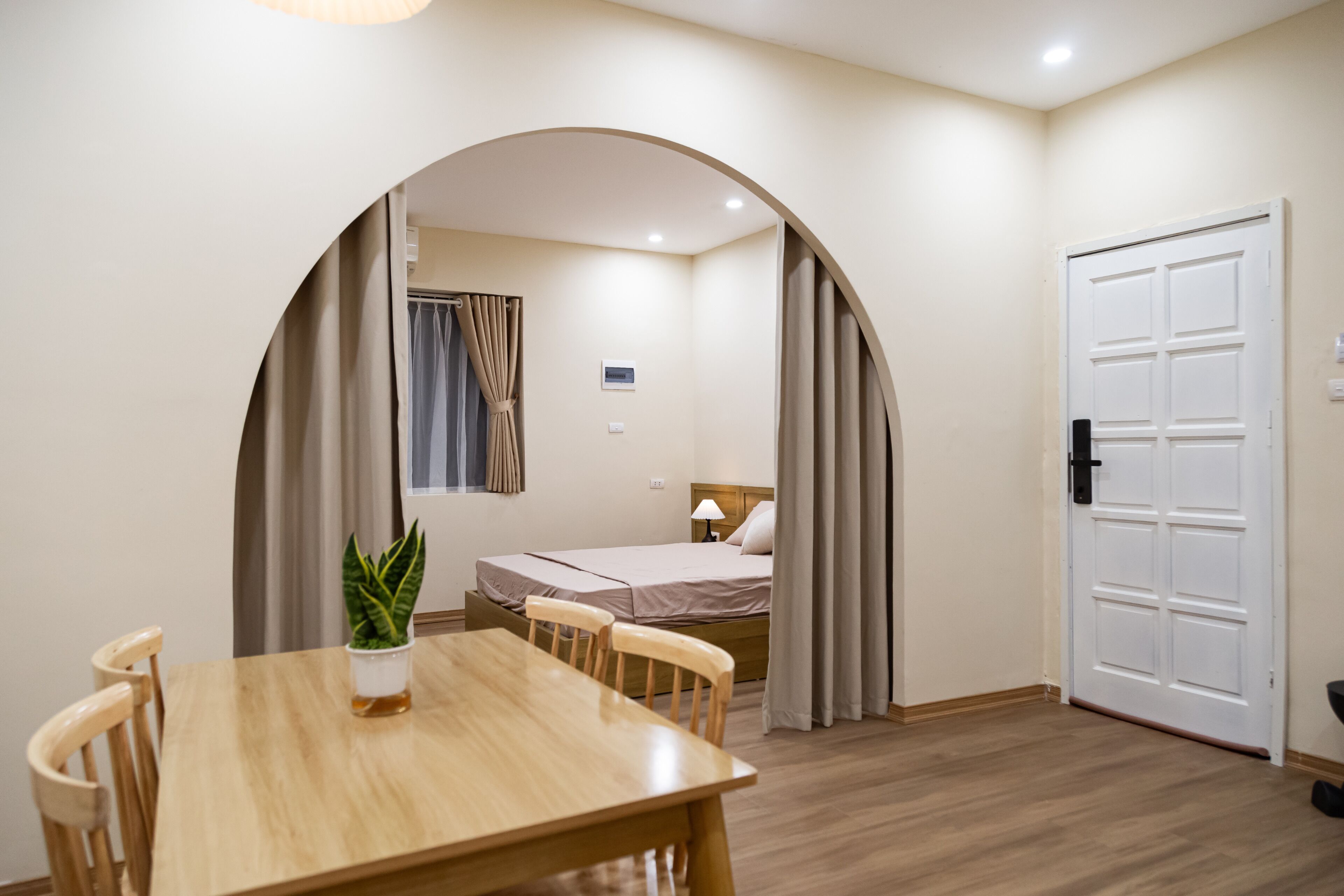 A room with a wooden dining table, two chairs, a bed with a headboard, a white door, and a small potted plant.