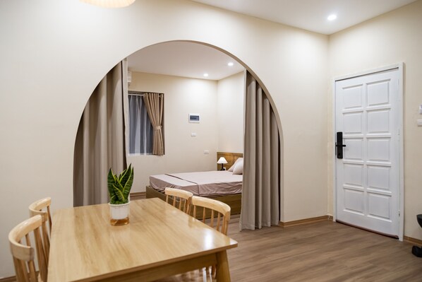 A room with a wooden dining table, two chairs, a bed with a headboard, a white door, and a small potted plant.