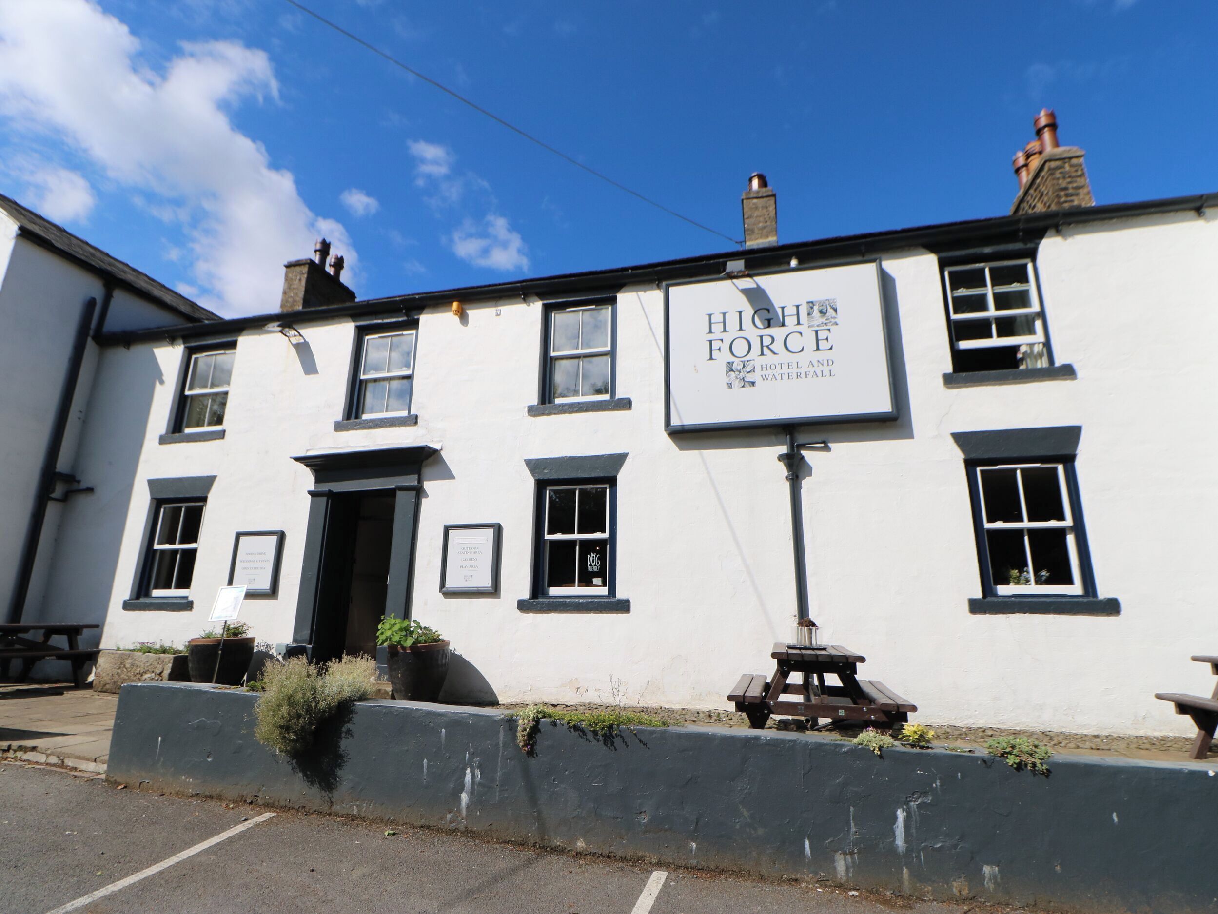 A two-story building with a sign reading "High Force Hotel and Waterfall". There are multiple windows, a door, and a small outdoor seating area with a table and chairs.