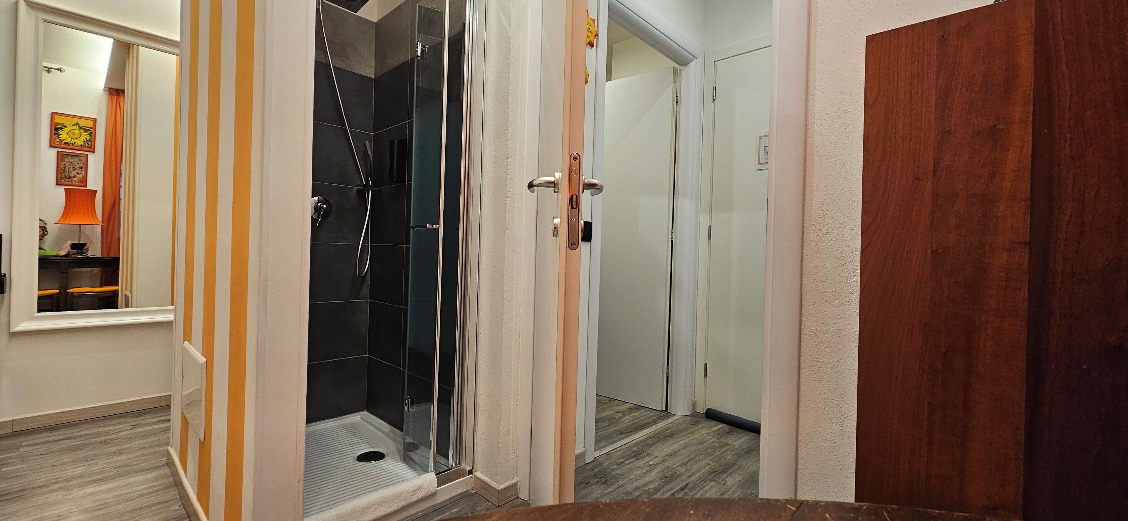Un pasillo con cabina de ducha de vidrio, una puerta de madera y un espejo que refleja una habitación con una lámpara y una silla.