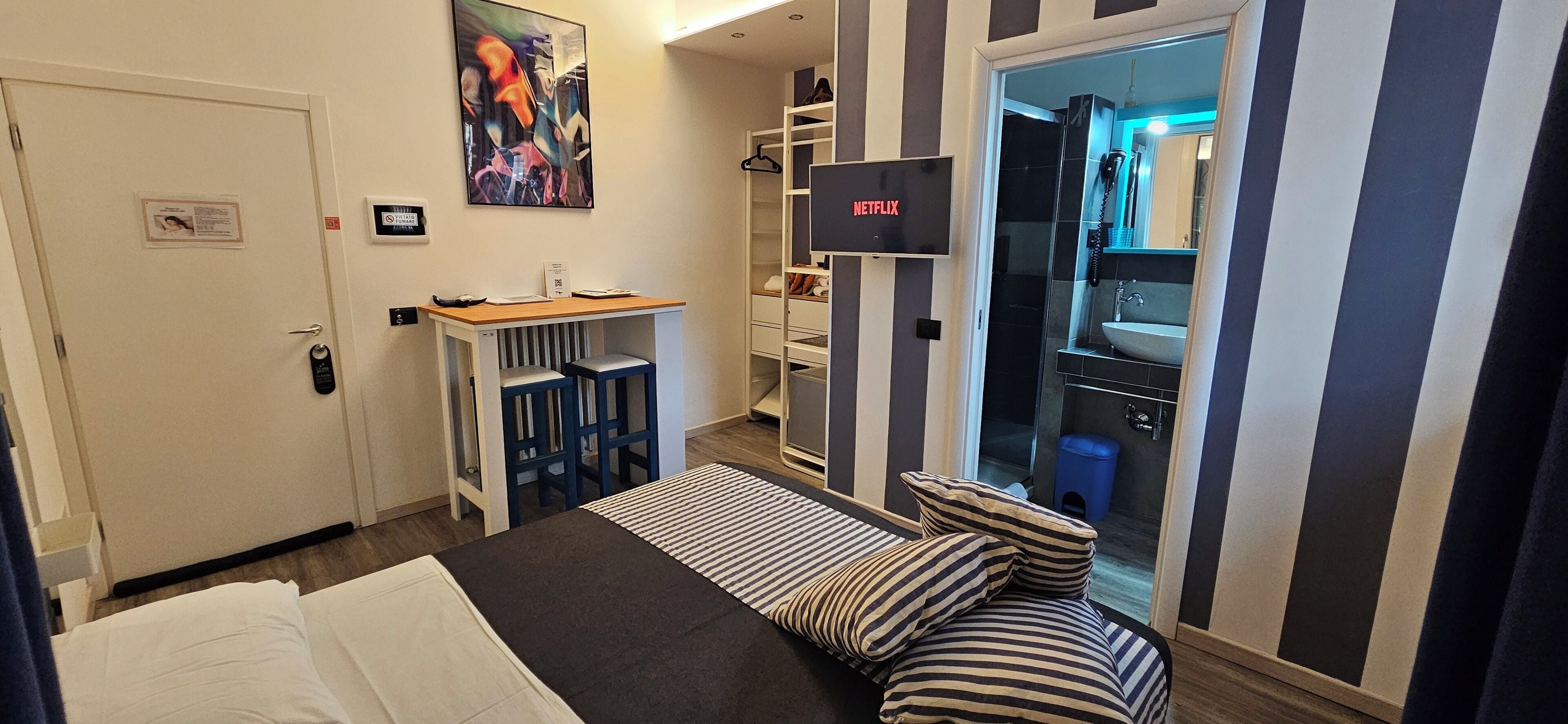 Ein Hotelzimmer mit Bett, Schreibtisch mit Stuhl, Wandfernseher, Badezimmer mit Waschbecken und Spiegel sowie Streifenmuster an den Wänden.