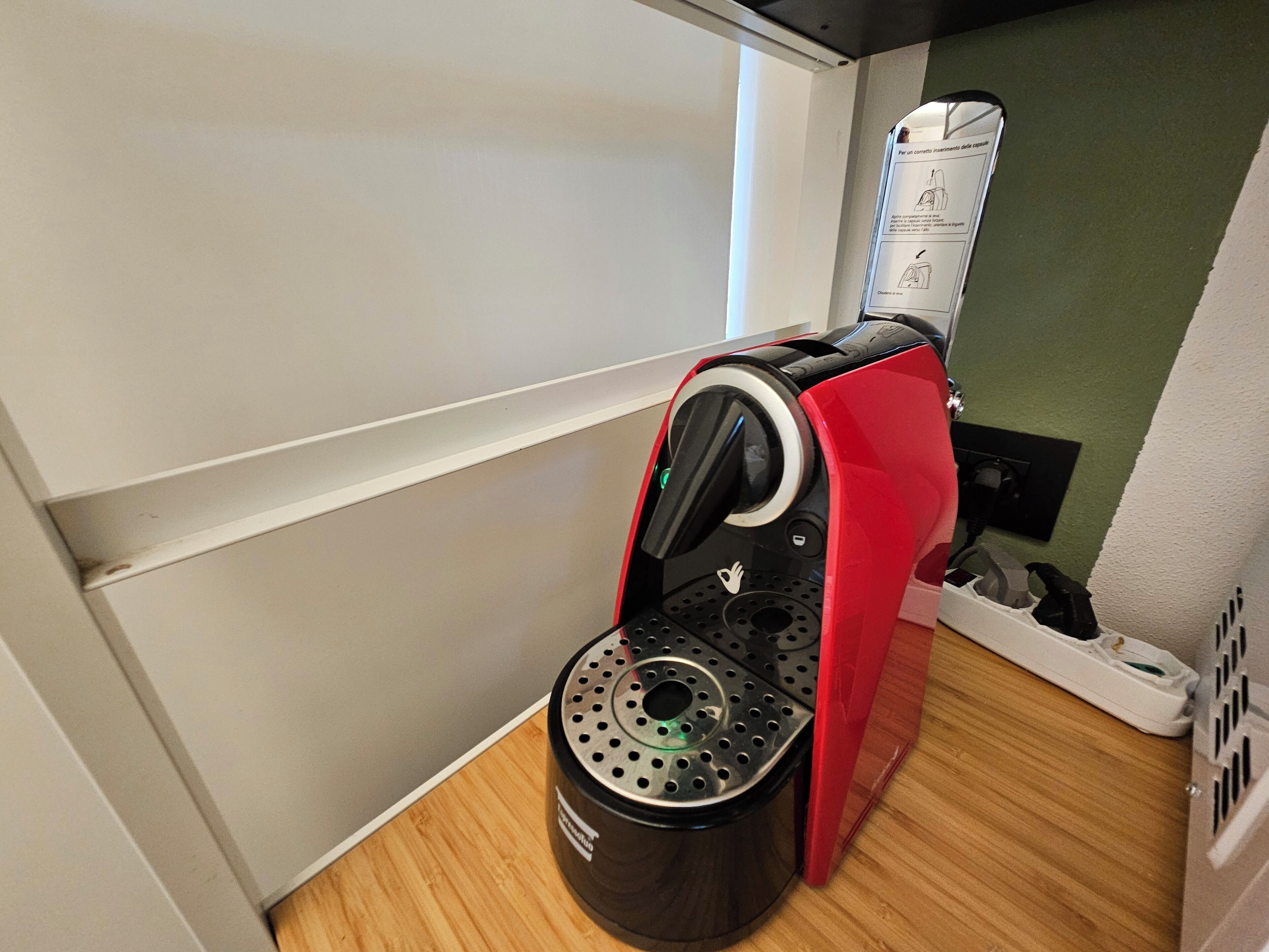 A compact coffee machine with a red and black design, placed on a wooden surface in a room with white walls and a green accent wall.