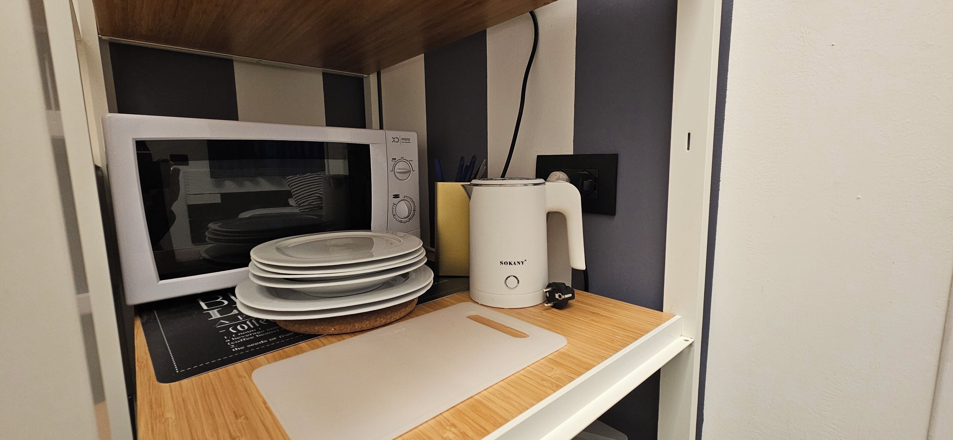 Une petite cuisine équipée d’un four à micro-ondes, d’une bouilloire et d’une pile d’assiettes posées sur un plan de travail en bois.
