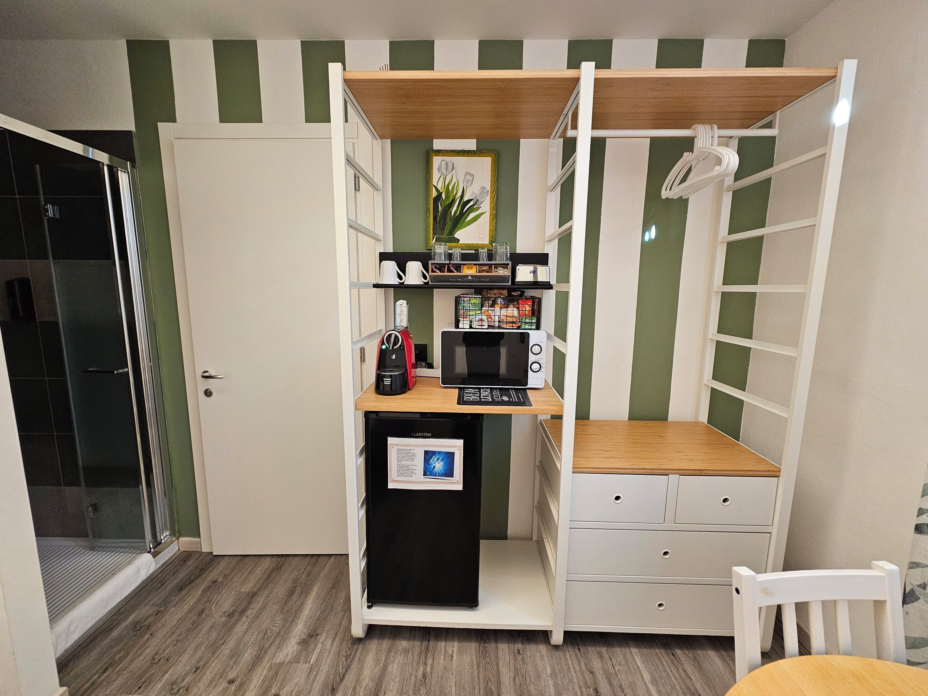 Una kitchenette compacta con microondas, cafetera y refrigerador pequeño. Hay un estante de madera sobre la encimera y una silla blanca a la derecha.