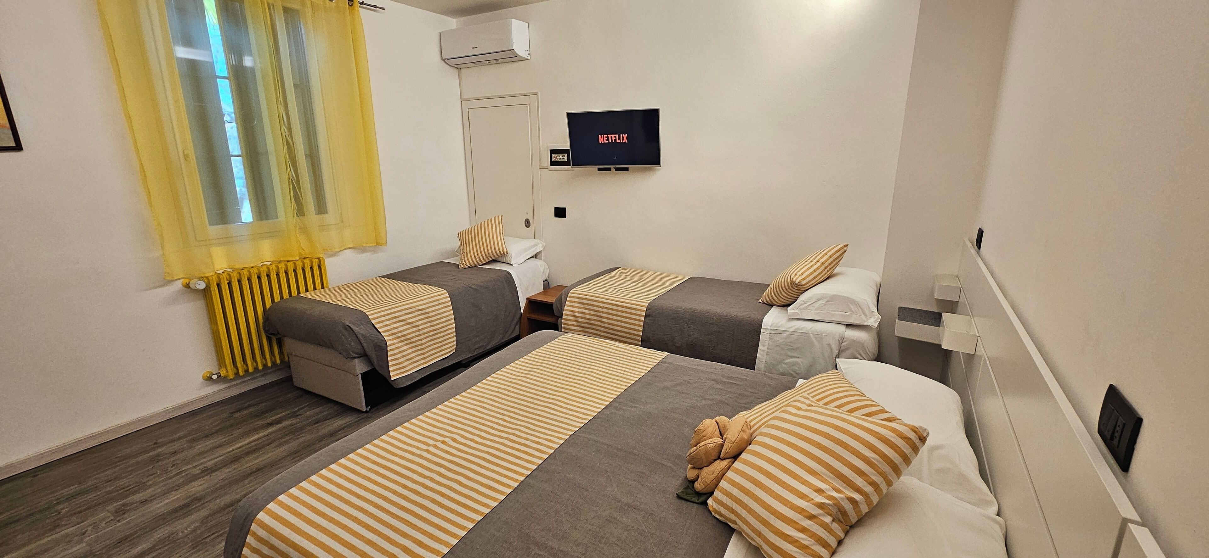 Camera d'albergo con due letti, una TV fissata al muro e una finestra con tende gialle.