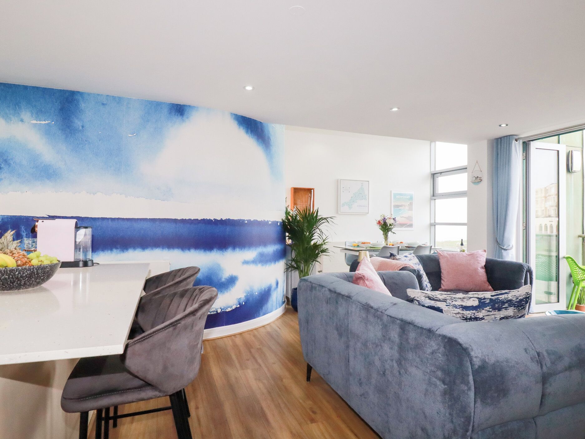 A modern living room with a large blue abstract painting, a grey sofa, a dining area with a white table, and a view through large windows.