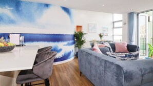 Ett modernt vardagsrum med en stor, blå, abstrakt målning, en grå soffa, en matplats med ett vitt bord och utsikt genom stora fönster.