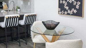 Een moderne keuken met donkere kastjes, een wit aanrechtblad en een zwart-wit bloemschilderij. Er staan twee barkrukken met gestreepte kussens en een ronde glazen tafel met een zwarte kom.