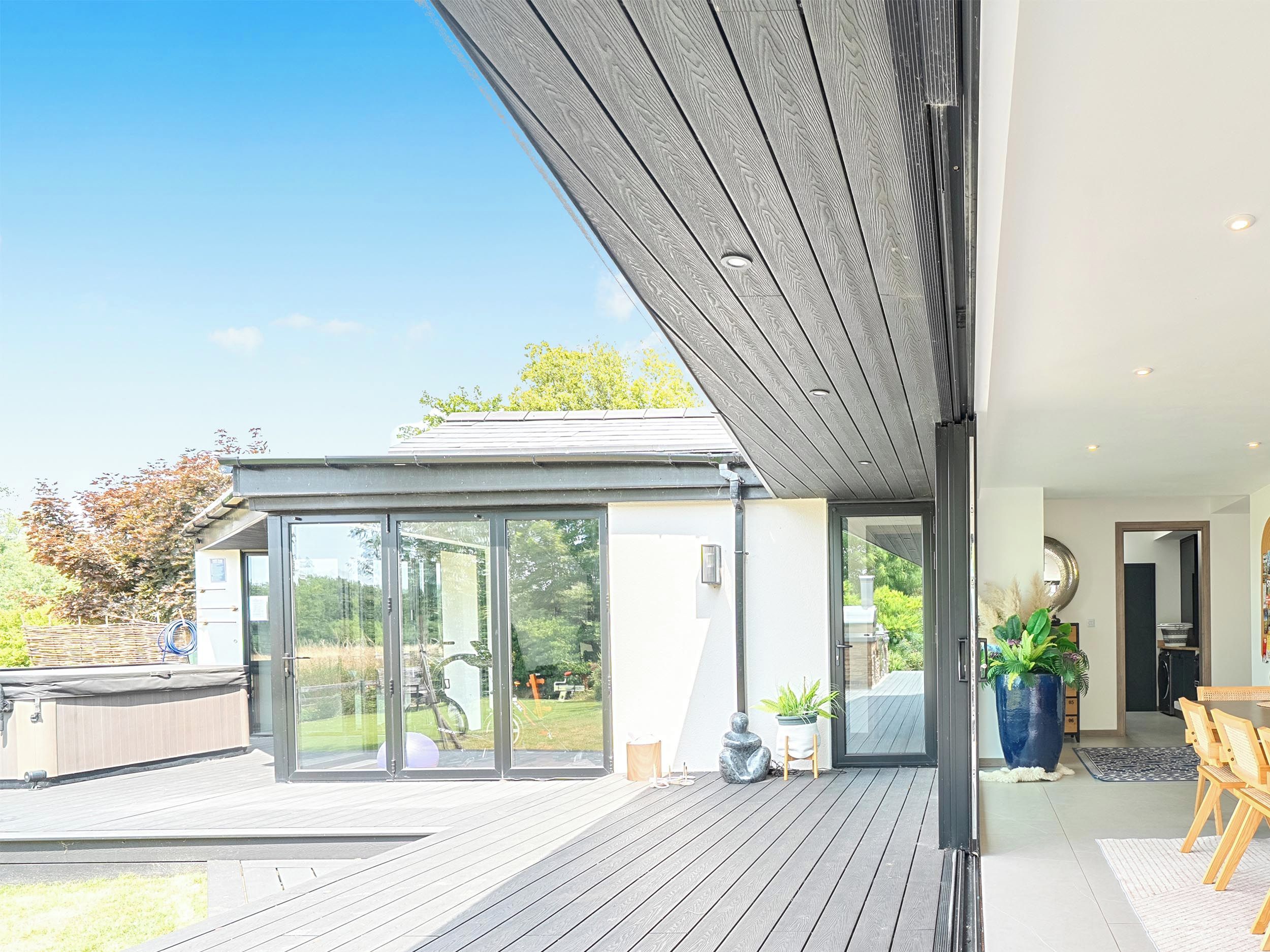 Una casa moderna con una terrazza in legno, ampie porte scorrevoli in vetro e un'area pranzo con tavolo e sedie.