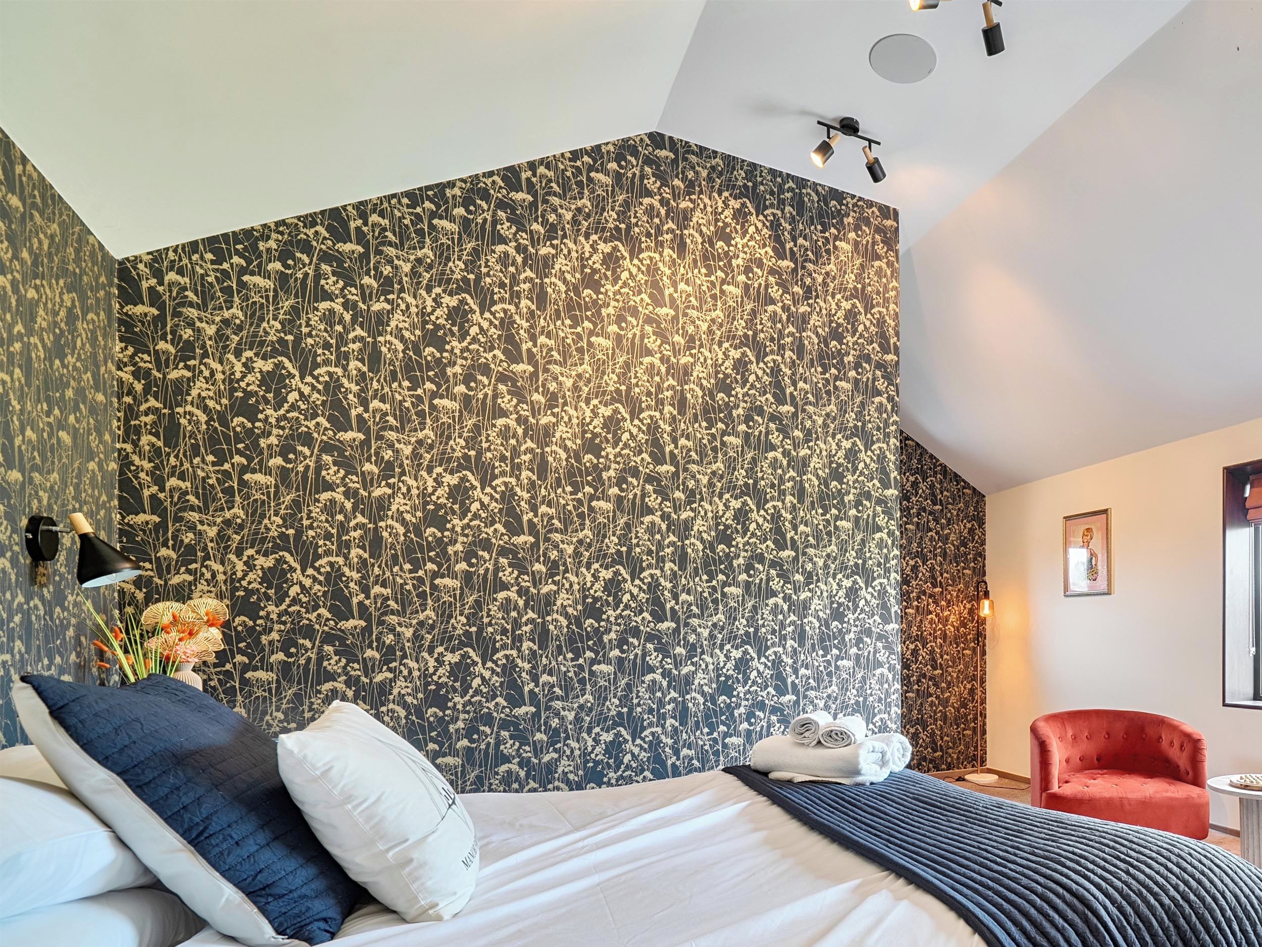 A modern hotel room with a large bed, a red armchair, and a wall with a floral pattern.