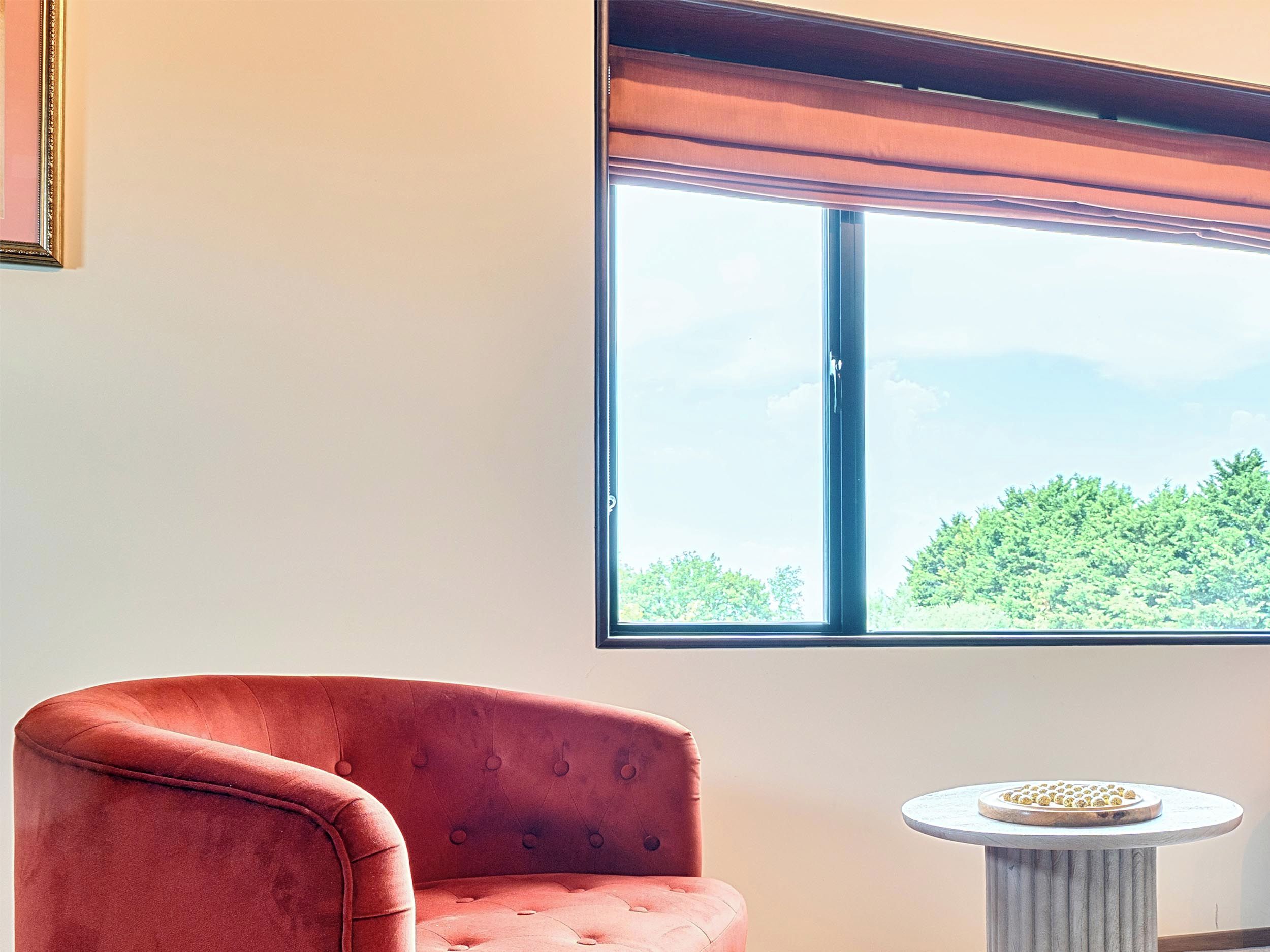 A room with a large window, a red upholstered chair, and a small round table with a decorative item.