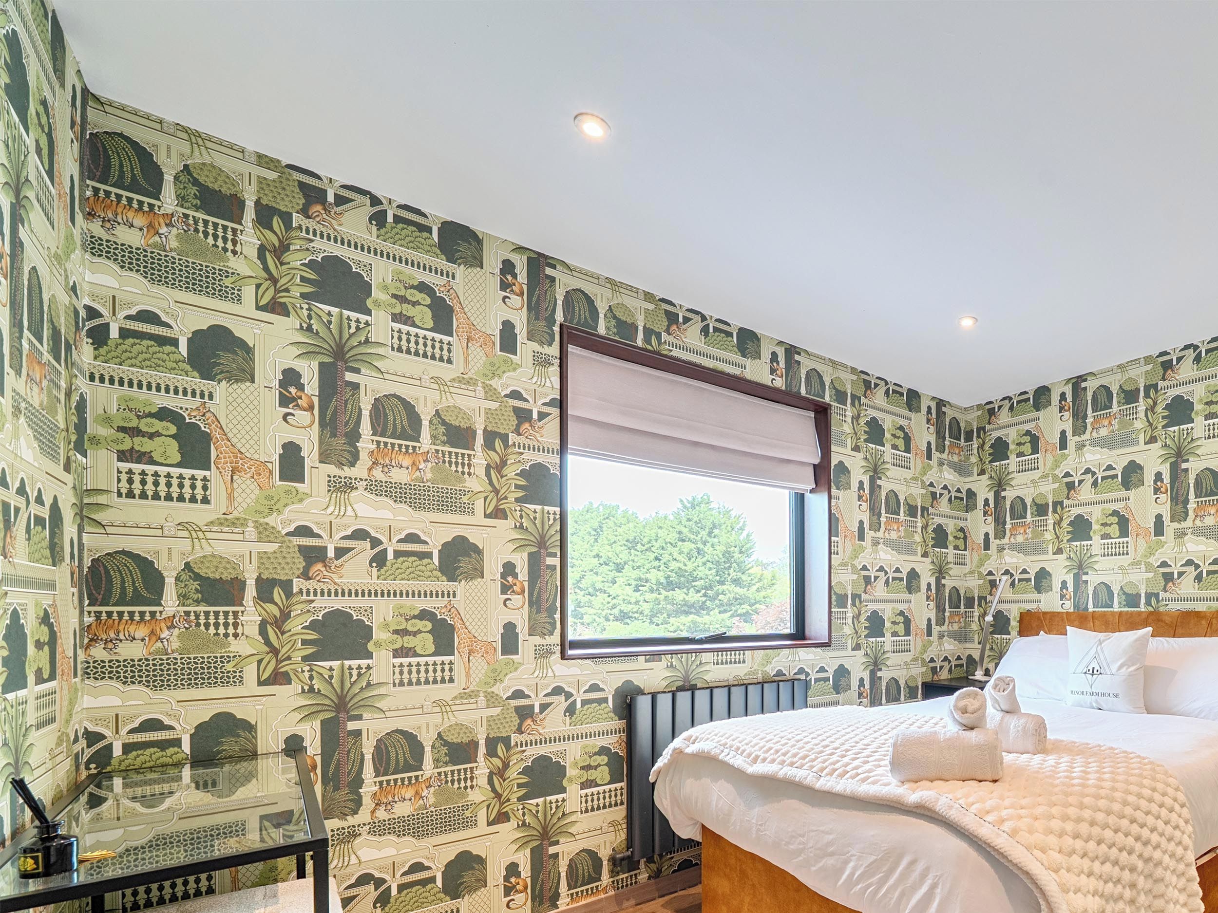 A bedroom with a large window, a bed with white bedding, and a wall with a nature-themed wallpaper.