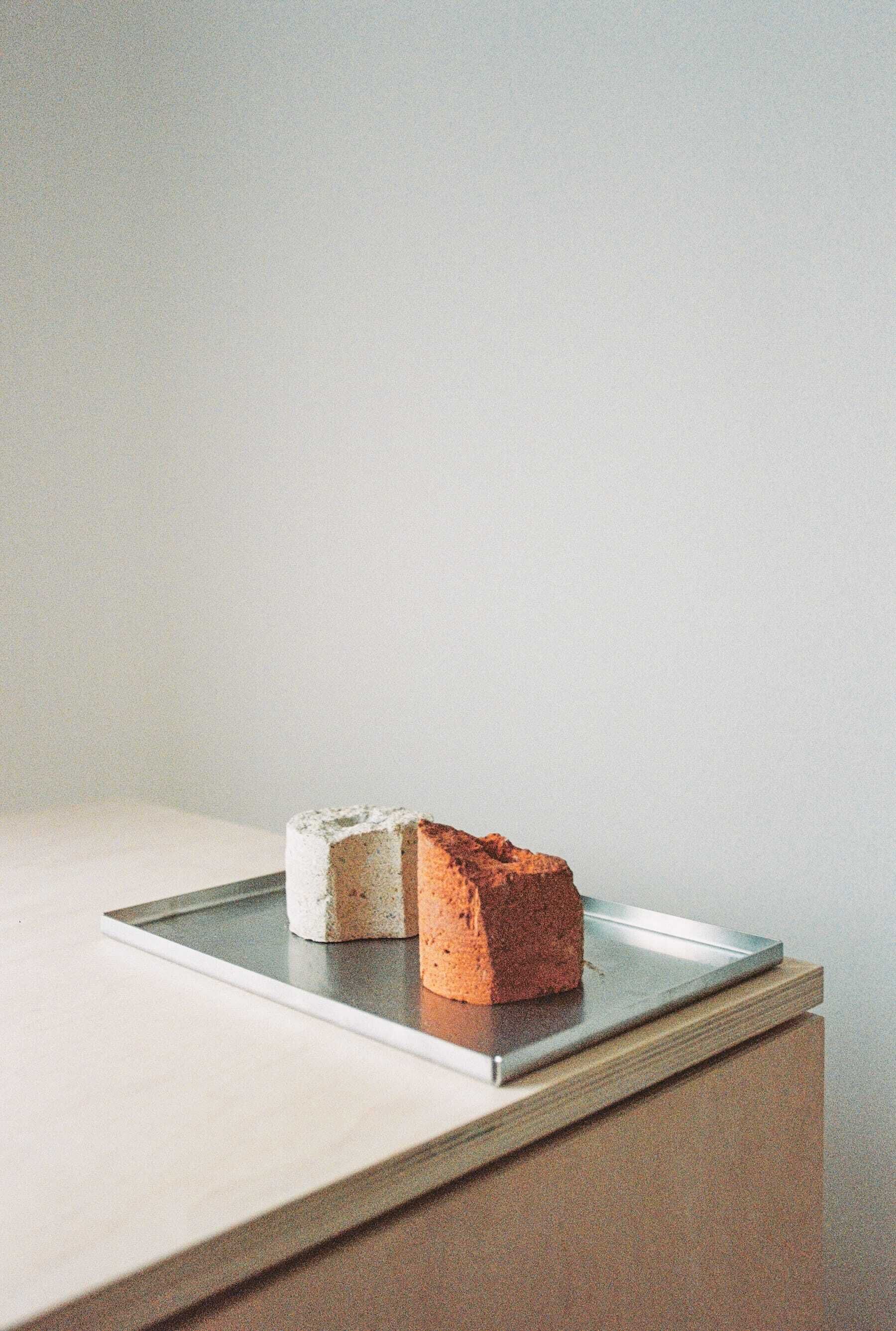 Une table minimaliste composée de deux blocs de matière, l’un blanc et l’autre d’une couleur entre le rouge et le brun, posés sur une surface réfléchissante.