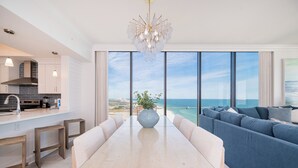 Une cuisine moderne dotée d’un grand îlot central, un coin repas avec une table en verre et un salon offrant une vue sur l’océan.