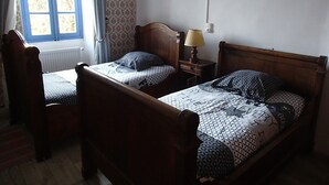 A room with two wooden beds, a nightstand with a lamp, a window with curtains, and a radiator. - Sabatier Apartment at the Eco-Museum – Authentic 1960s Farmhouse (Gissac)