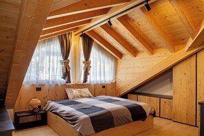 Une chambre avec un plafond en pente, des murs en bois, un lit recouvert d’une couverture à carreaux, une table de chevet avec une lampe et une fenêtre avec des rideaux.
