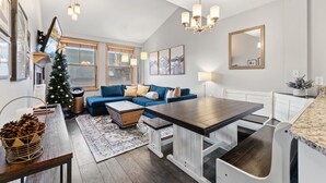 Uma sala de estar moderna com um sofá azul, mesa de centro de madeira e uma mesa de jantar com bancos. Uma árvore de Natal é visível no canto.
