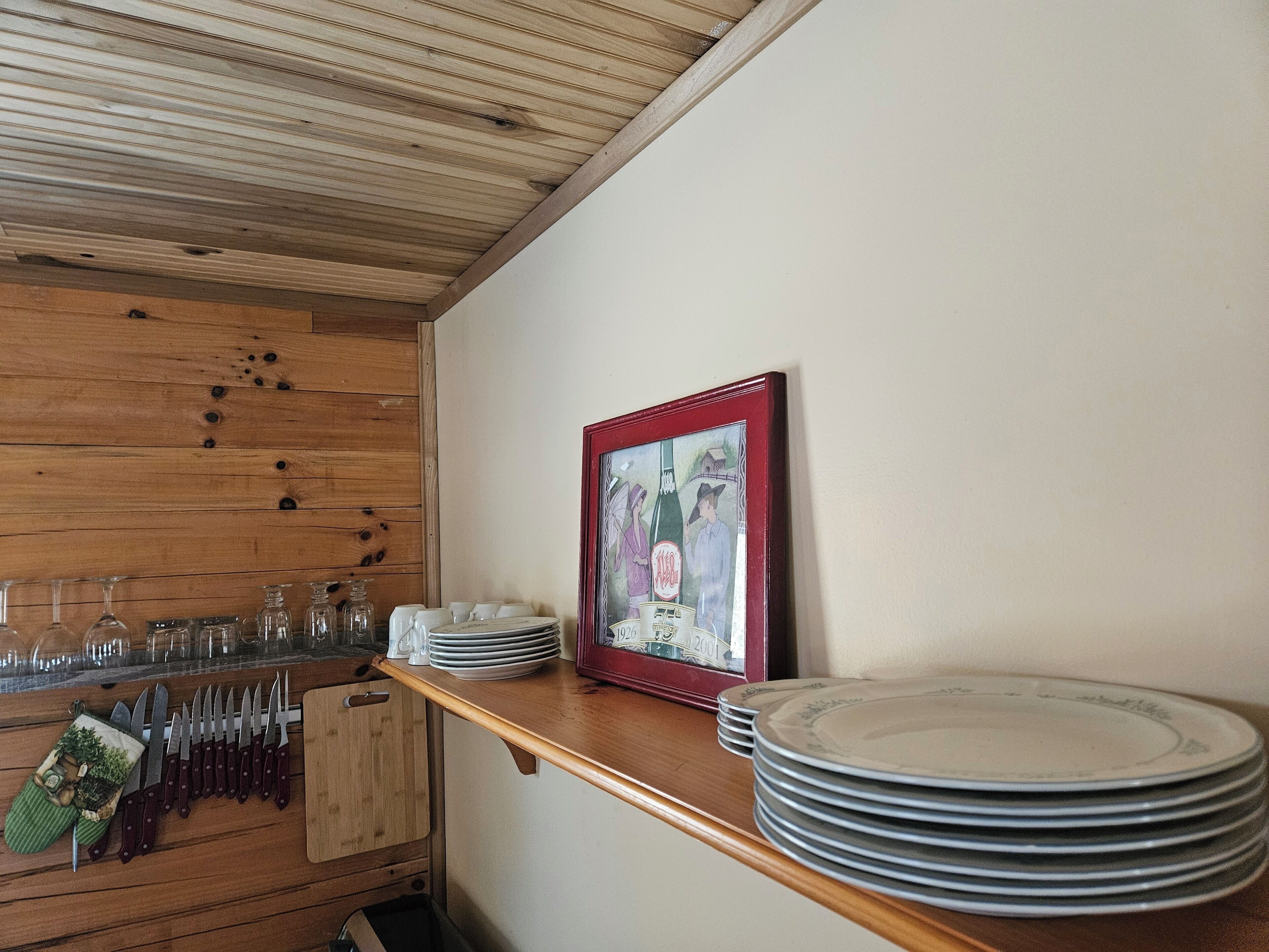 A kitchen with wooden cabinets, a wooden ceiling, a framed picture on the wall, and a counter with stacked plates and cutlery.