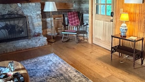 Ein Holzhütten-Interieur mit Kamin aus Stein, Holzwänden, einem Schaukelstuhl, einem Tisch mit Lampe und einer Tür mit Fenster.