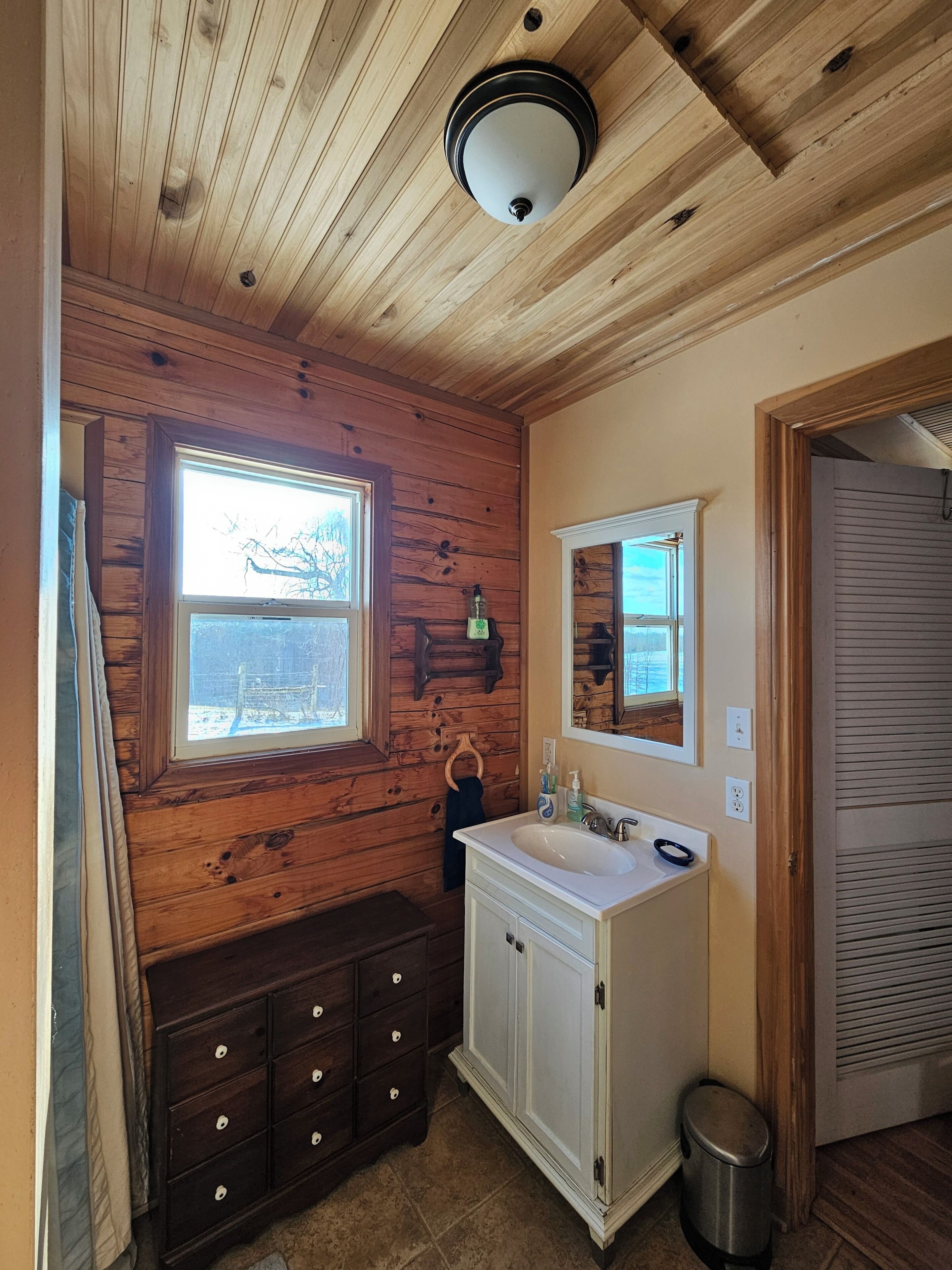 Une salle de bain aux murs lambrissés, équipée d’un lavabo, d’un miroir et d’une fenêtre.