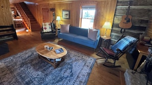 Ein gemütliches Zimmer mit Holzbalkendecke, einem blauen Sofa, einem Schaukelstuhl, einem Couchtisch, einer Gitarre und einer Treppe, die in einen weiteren Bereich führt.