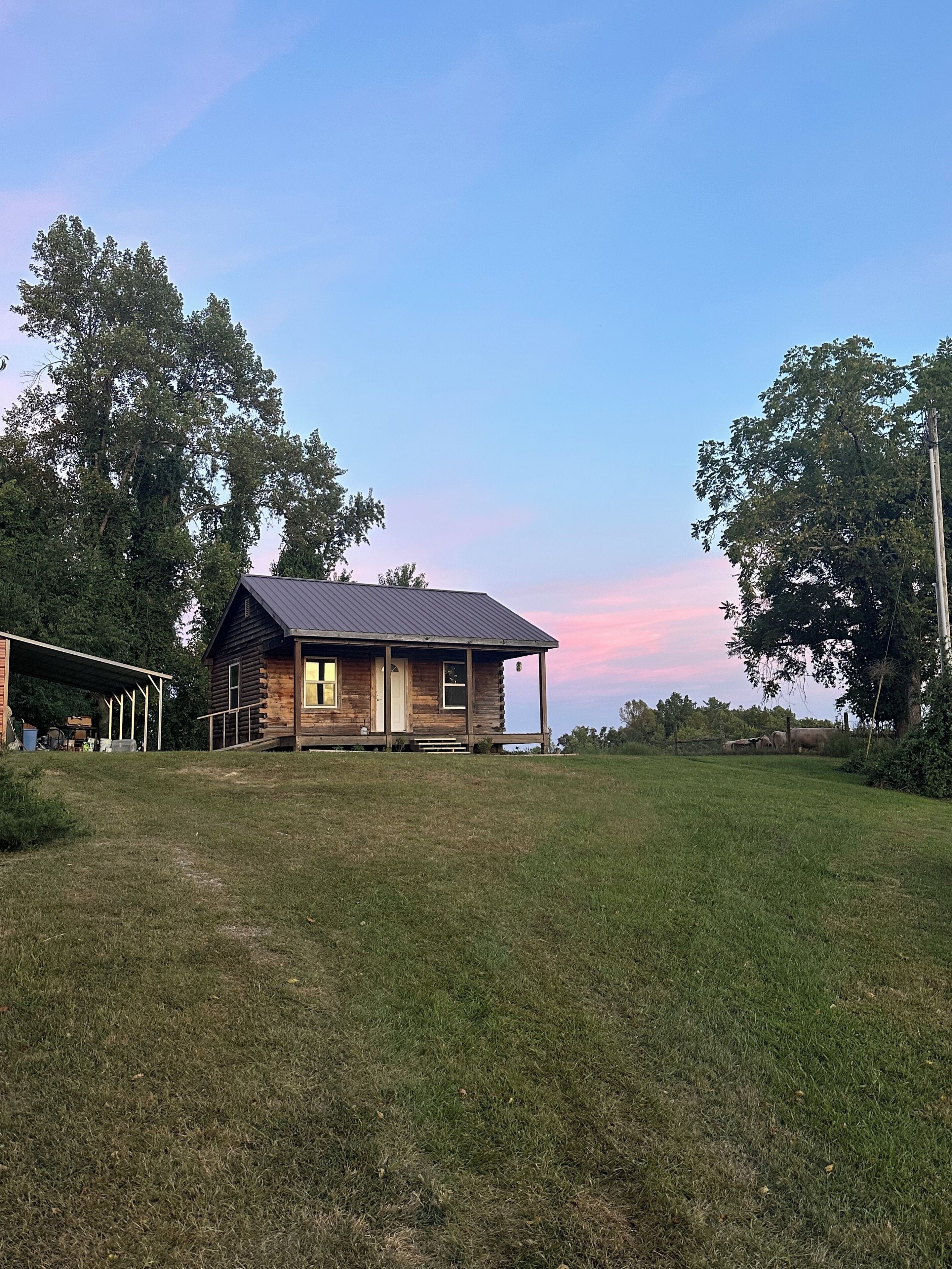 A rustic log cabin with a covered porch, surrounded by a grassy yard and trees.