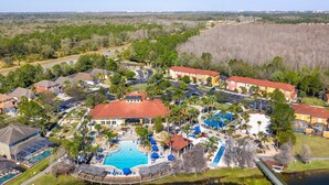 An aerial view of a resort with a central pool, multiple buildings, and surrounding greenery.