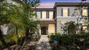 A two-story house with a covered entrance, surrounded by lush greenery and a paved pathway leading to the door. - Bella Vida Cozy Home | 15 min to Disney | Resort Style (Kissimmee)