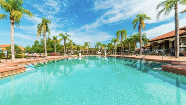 A clear swimming pool surrounded by palm trees and lounge chairs, with a building in the background. - Bella Vida Cozy Home | 15 min to Disney | Resort Style (Kissimmee)