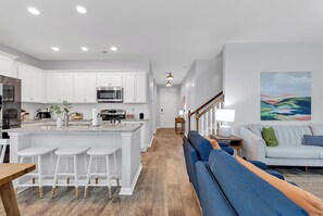 Une cuisine moderne avec des meubles blancs, un îlot central doté de tabourets, un coin repas avec une table en bois et un salon avec un canapé et un tableau au mur.