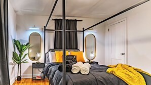 A four-poster bed with a black frame and a canopy, a bedside table with a lamp, a potted plant, and a mirror on the wall. - Limited time Deal! Cozy Luxe Escape Modern 2BR, sleeps 4,Couples, Business & Pet (Raleigh)