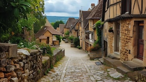 Uma rua estreita de paralelepípedos, ladeada por casas de pedra tradicionais, com uma parede de pedra de um lado e vista para colinas ao longe.